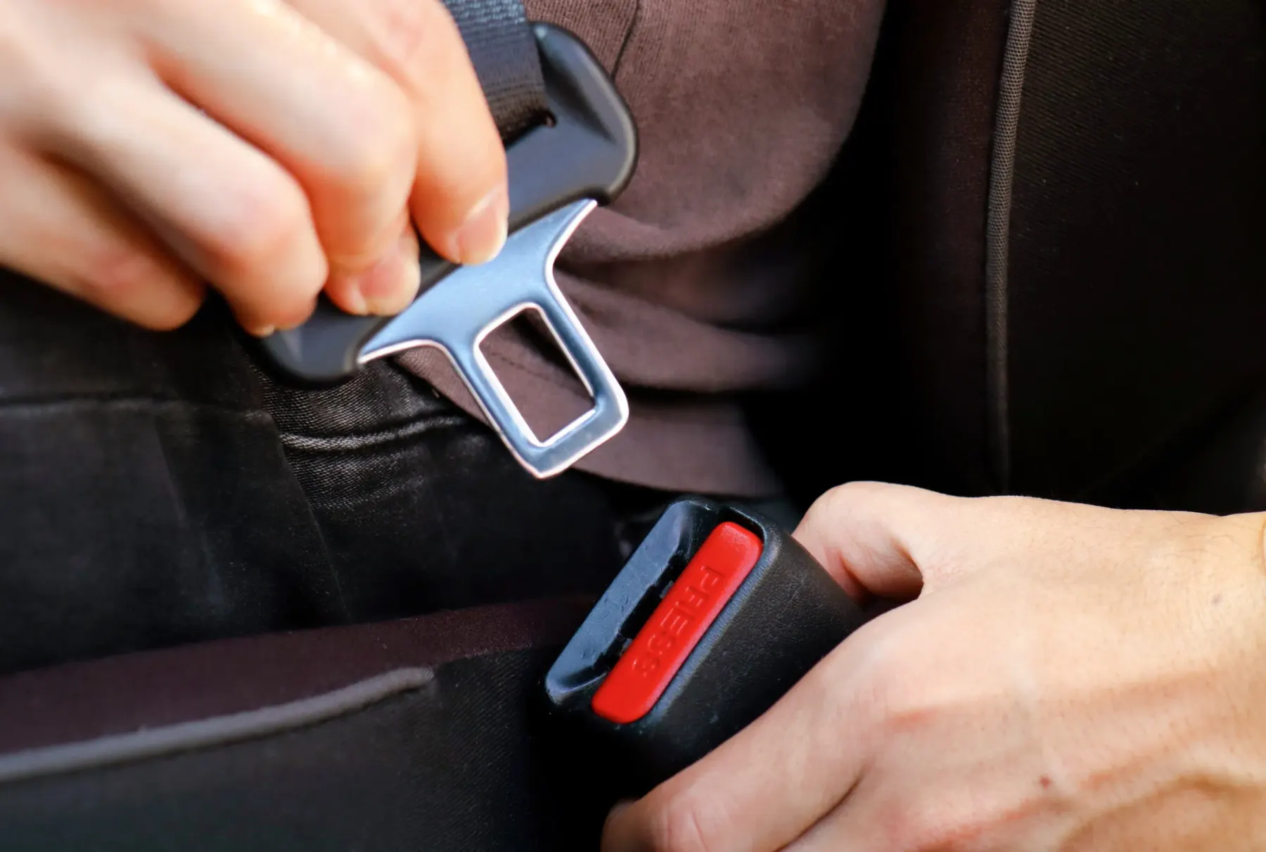 Two hands guiding a metal seat belt latch plate towards a black buckle receptacle with a red release button inside a vehicle