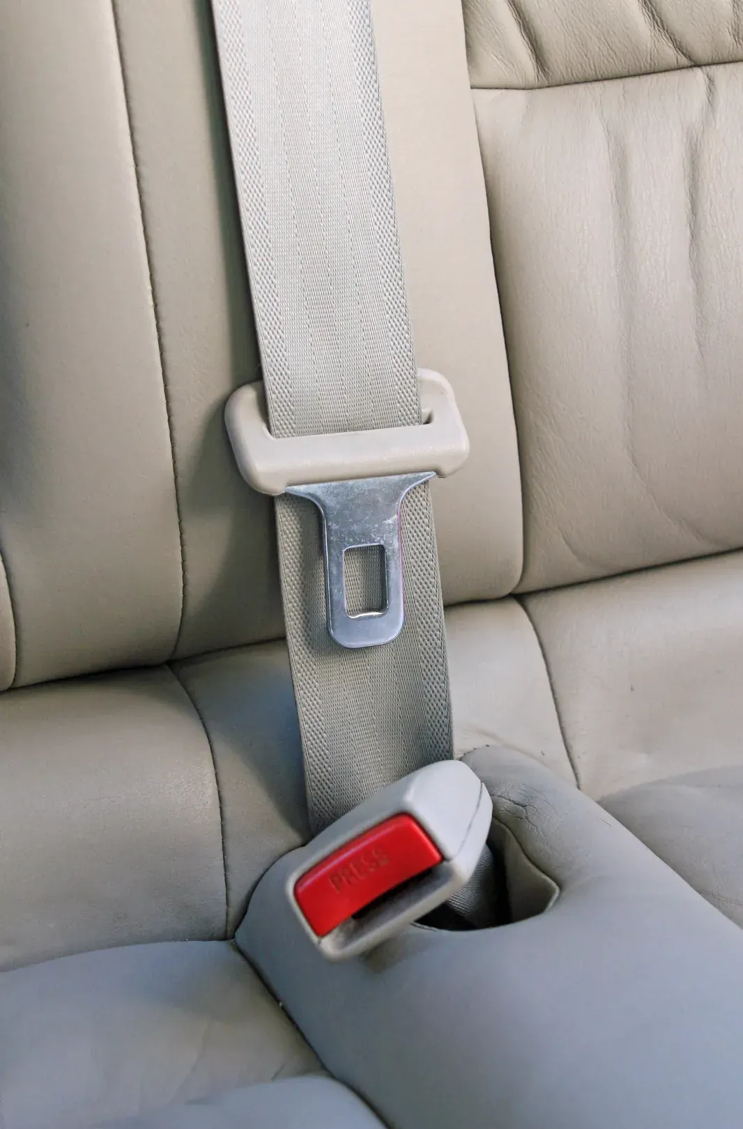 A light grey rear seat belt running through a moulded guide with its latch plate and a buckle marked PRESS on a cream leather rear bench