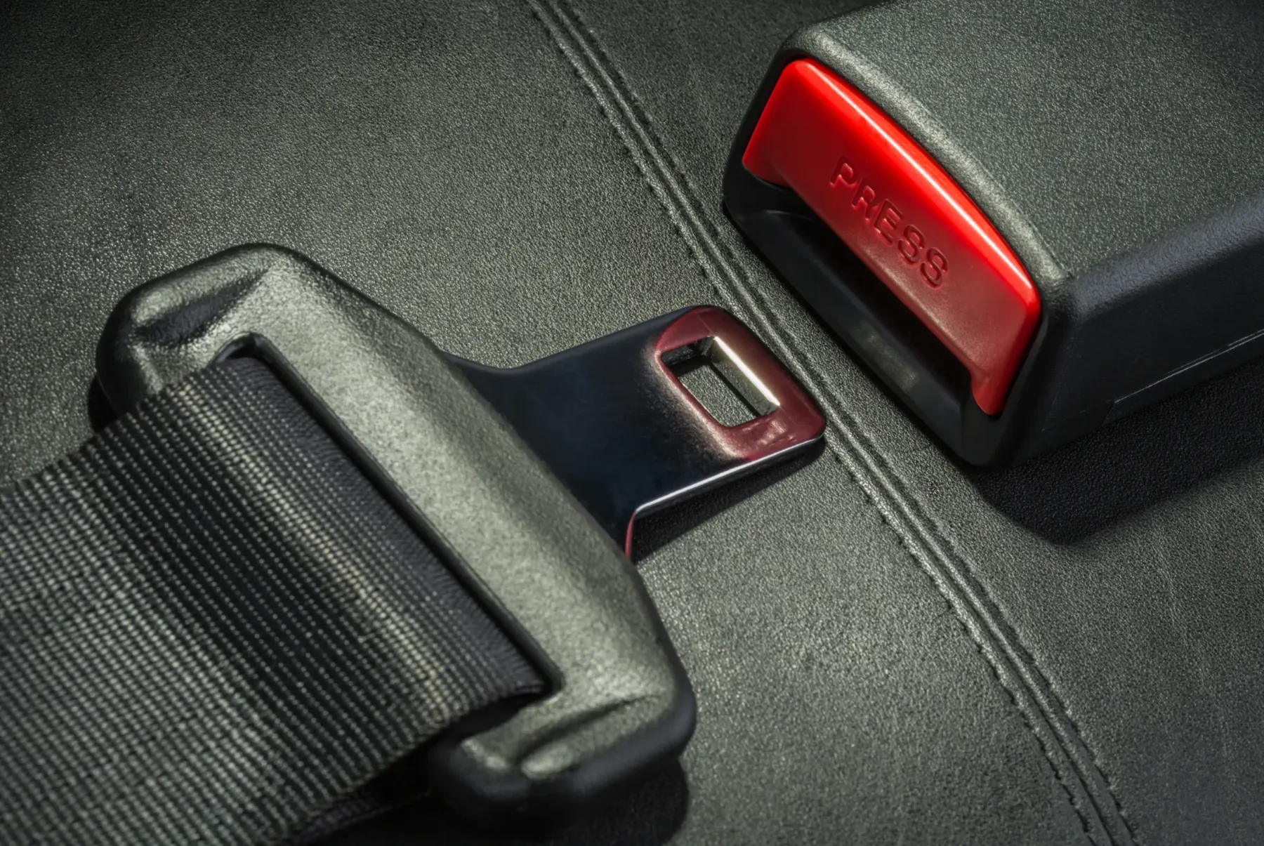 A three-point seat belt latch plate lying beside its matching black buckle receptacle with a red release button, photographed on a dark textured seat
